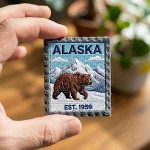 May include: Embroidered patch featuring the word "ALASKA" above a brown bear walking in front of snow-capped mountains. The patch has a blue sky background with white clouds and the text "EST. 1959". The patch is framed with a gray border.