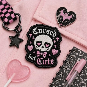 May include: A collection of gothic-themed accessories on a pink background. Includes a black star charm, a black and pink checkered strap, a heart-shaped lollipop, a black patch with a skull and the words "Cursed but Cute", and a heart-shaped bat pin.