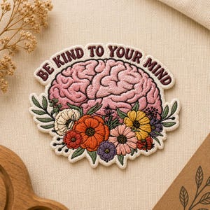 May include: Embroidered patch featuring a pink brain surrounded by colorful flowers. The patch has the text "BE KIND TO YOUR MIND" in a curved, dark red font. The flowers include orange, yellow, and purple blooms with green leaves.