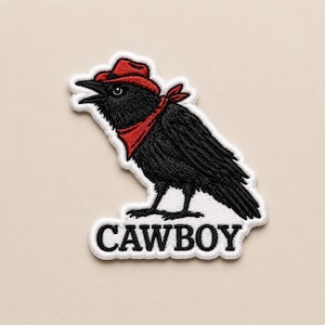 May include: An embroidered patch featuring a black crow wearing a red cowboy hat and a red bandana around its neck. The crow is depicted in profile facing left. Below the crow, the word CAWBOY is embroidered in black capital letters on a white background.