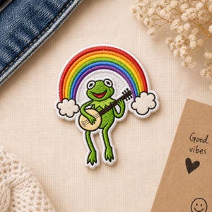 May include: Embroidered patch featuring a green frog playing a banjo under a rainbow. The rainbow has red, orange, yellow, green, blue, and purple stripes. White clouds frame the rainbow. The patch is on a beige background.