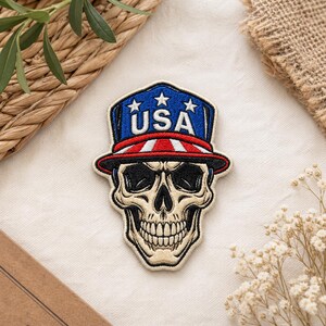 May include: Embroidered patch featuring a skull wearing a patriotic hat. The hat is blue with white stars and the word "USA". The brim has red and white stripes. The skull is beige with black eye sockets and teeth.