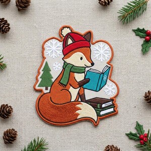 May include: An embroidered patch featuring a fox with orange fur and a cream-colored chest, wearing a red winter hat and a green scarf. The fox is reading a blue book, with a green pine tree, white snowflakes, and stacked books nearby.
