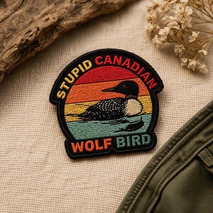 May include: Embroidered patch with a retro sunset design featuring a loon and the text "STUPID CANADIAN WOLF BIRD". The patch has a black border and is likely for jackets, bags, or other apparel.