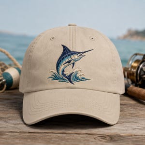 May include: A tan baseball cap featuring an embroidered marlin leaping from ocean waves. The marlin is blue and white, with detailed stitching. The cap has a curved brim and an adjustable strap.