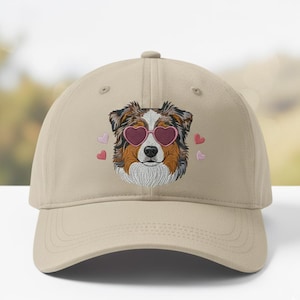 May include: Tan baseball cap featuring an embroidered dog wearing heart-shaped sunglasses. The dog has brown, white, and black fur, with pink hearts embroidered around it. A stylish accessory for dog lovers.