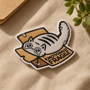 May include: Embroidered patch featuring a cartoon cat inside a cardboard box. The cat is gray and white with black stripes. The patch has the words "Emotionally Fragile" in gold lettering.
