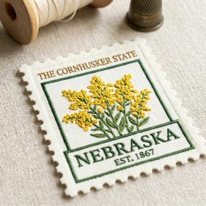 May include: Embroidered patch in the shape of a postage stamp featuring yellow flowers and green leaves. The text "NEBRASKA EST. 1867" is displayed. The words "THE CORNHUSKER STATE" are at the top. A wooden spool and thimble are in the background.