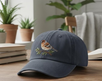 May include: A navy blue baseball cap featuring an embroidered bird perched on a branch. The bird has brown, white, and black feathers. The cap is made of a soft fabric and has a curved brim.