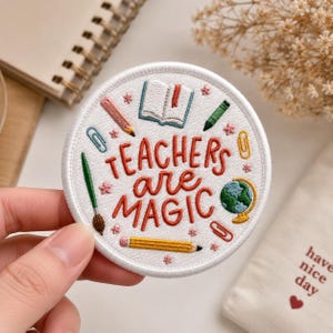 May include: A circular white embroidered patch featuring the text "TEACHERS are MAGIC" in red thread. The design includes colorful stitched icons of an open book, a globe, a paintbrush, a yellow pencil, a pink pencil, and paper clips.