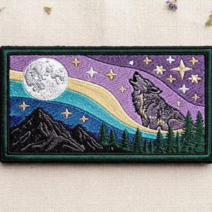 May include: A rectangular embroidered patch featuring a howling wolf silhouette against a purple night sky with a large white moon, golden stars, a flowing blue and gold aurora, dark mountain peaks, and a line of green pine trees at the base.