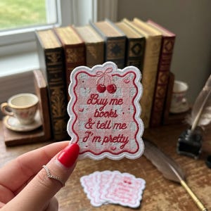 May include: A pink and white embroidered patch with the text "Buy me books & tell me I'm pretty" and cherry illustrations. The patch is held in front of a collection of vintage books and a teacup.
