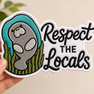May include: An embroidered patch featuring a grey manatee swimming in teal water with green seagrass. The patch includes the black embroidered text "Respect THE Locals" on a white background with a white border and a die-cut shape.