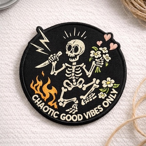 May include: Circular black patch with a skeleton design. The skeleton holds a knife and flowers, with a lightning bolt and flames. Text reads "CHAOTIC GOOD VIBES ONLY". The skeleton is cream, flames are orange.