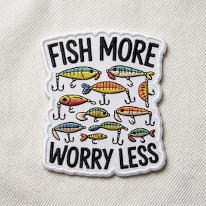 May include: A white embroidered patch with black text that reads FISH MORE WORRY LESS. The design features various colorful fishing lures in shades of yellow, blue, orange, and green, each equipped with metal-colored hooks, arranged on a white background.