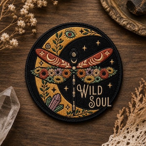 Peut inclure: Écusson brodé avec un motif de libellule, des ailes florales, un croissant de lune et le texte "WILD SOUL". L'écusson a un fond noir avec des accents dorés, orange, rouges et verts. Des étoiles et des cristaux sont également inclus dans le design.