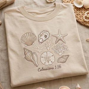 May include: Beige t-shirt featuring an embroidered design of various seashells, a starfish, and coral. The text "Colossians 1:16" is embroidered below the design. The shirt is folded and displayed on a neutral surface.