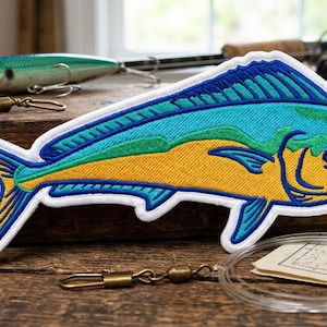 May include: An embroidered patch shaped like a mahi-mahi fish, featuring vibrant yellow, bright blue, and green thread colors with a white border. The fish design displays a detailed fin structure and a distinct eye, rendered in a flat, stitched style.
