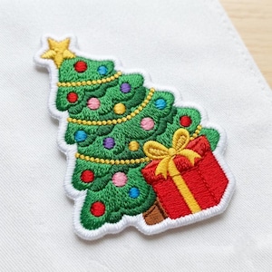 May include: An embroidered patch featuring a green Christmas tree topped with a yellow star. The tree is decorated with yellow garland and multi-colored ornaments in red, blue, pink, and purple. A red gift box with a yellow bow sits at the base.