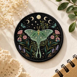 May include: Circular embroidered patch with a Luna moth design. The moth is green with crescent moon details, set against a black backdrop with pink flowers, butterflies, and foliage. Ideal for customising clothing or accessories.