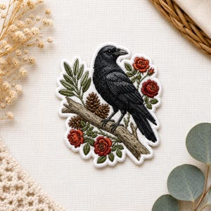 May include: Embroidered patch depicting a black crow on a branch, surrounded by red roses, pine cones, and green leaves. The patch has a white border, suitable for application on fabric items.