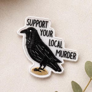 May include: Embroidered patch featuring a black crow with the text "SUPPORT YOUR LOCAL MURDER" in black lettering on a white background. The crow is perched on a gold-colored base, with a white border.