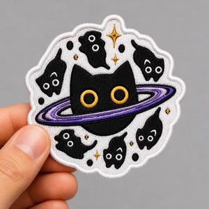 May include: An embroidered patch featuring a large black cat head centered with a purple planetary ring. The design includes smaller black cat silhouettes, gold stars, and black dots on a white background with a scalloped white border edge.