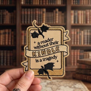 May include: Embroidered patch with a beige background and a gold border. The patch features the text "a reader without their KINDLE is a tragedy" with black dragon silhouettes. The patch is held in a hand.