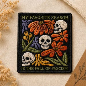May include: A rectangular embroidered patch with a black background featuring three white skulls, orange and yellow flowers, and green leaves. The text MY FAVORITE SEASON IS THE FALL OF FASCISM is embroidered in yellow at the top and bottom.