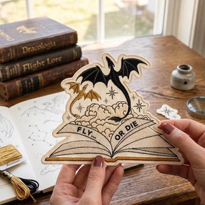 May include: Embroidered patch depicting a black dragon and a gold dragon above an open book with the text "FLY... OR DIE". The patch has a cream background with embroidered clouds and stars. The patch is held in front of a wooden table.