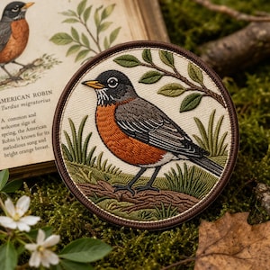 May include: Embroidered patch featuring an American Robin. The bird has a bright orange breast, gray back, and black head. The patch is round with a brown border and includes a sprig of leaves and grass. Text on the left reads "AMERICAN ROBIN Turdus migratorius".