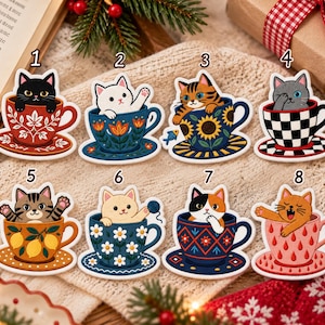 May include: Eight die-cut stickers featuring cats in teacups. 1: black cat in red floral cup. 2: white cat in blue floral cup. 3: tabby cat in sunflower cup. 4: gray cat in checkered cup. 5: tabby cat in lemon cup. 6: cream cat in daisy cup. 7: calico cat in geometric cup. 8: orange cat in pink raindrop cup.