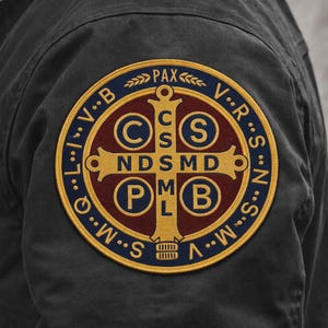 May include: A circular embroidered patch featuring a Saint Benedict cross design in gold, dark red, and navy blue. The emblem displays the letters C S S M L N D S M D, C S P B, and PAX, with a navy blue border containing the letters V R S N S M V S M Q L I V B.