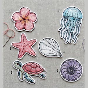 May include: Embroidered patches featuring sea life and floral designs. Includes a pink flower, a blue jellyfish, a pink starfish, a white shell, a sea turtle, and a purple nautilus shell. Each patch has a white border and is on a gray fabric background.