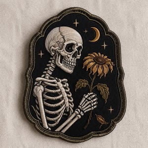 May include: Embroidered patch featuring a detailed skeleton holding a sunflower. The design is set against a black background with a decorative border, a crescent moon, and stars. The patch has a gothic or dark aesthetic.