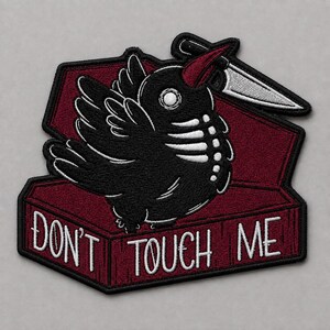 May include: An embroidered patch featuring a black bird with a white skeleton ribcage design, holding a silver knife in its beak. The bird is positioned inside a dark red coffin with the white text DON'T TOUCH ME printed on the front panel.