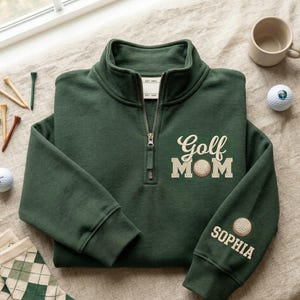 May include: A dark green quarter-zip sweatshirt with the embroidered text "Golf Mom" and a golf ball graphic. The sleeve features a golf ball graphic and the name "Sophia". Golf tees, a golf ball, and argyle socks are also visible.