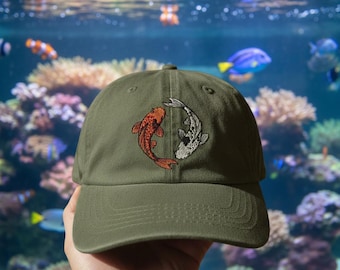 May include: Olive green baseball cap featuring an embroidered design of two koi fish, one orange and one white, in a yin-yang arrangement. The cap has a curved brim and a classic six-panel design, suitable for casual wear.