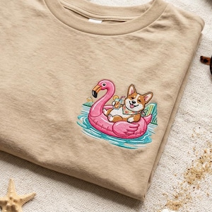 Puede incluir: Camiseta color beige con un diseño bordado de un perro corgi relajándose en un flotador de flamenco rosa en una piscina. El perro lleva un pañuelo y sostiene una bebida. Gafas de sol visibles en el fondo.