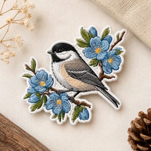 May include: Embroidered patch of a bird with a black head, grey and beige body, and a long tail. The bird is perched on a branch with blue flowers and green leaves. The patch has a white border.