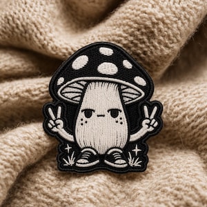 May include: Black and white mushroom patch with a peace sign gesture. The mushroom has a black cap with white spots and a beige stem. The patch is embroidered with a detailed design and is set against a beige knitted fabric.