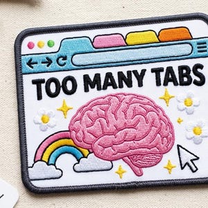 May include: A rectangular embroidered patch featuring a white background with a browser window graphic at the top, the black text "TOO MANY TABS," a central pink brain, a small rainbow with clouds, yellow stars, and white flowers with yellow centers.