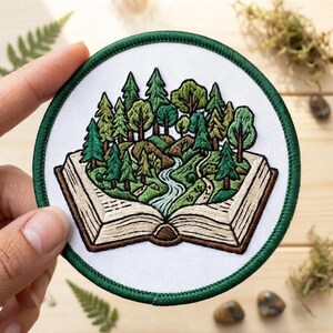 May include: A circular embroidered patch featuring an open book with a landscape emerging from its pages. The scene depicts a winding river flowing through a dense forest of green pine and deciduous trees, all set against a white background with a dark green border.