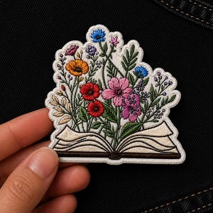 May include: Embroidered patch featuring a colorful bouquet of flowers bursting from an open book. The flowers include red, pink, orange, and blue blooms with green leaves. The patch has a white border and is designed for sewing or ironing onto fabric.