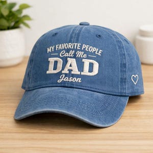 May include: A blue denim baseball cap with the text "MY FAVORITE PEOPLE Call Me DAD Jason" embroidered in white. A white heart is embroidered on the side of the cap. The cap is sitting on a wooden surface.