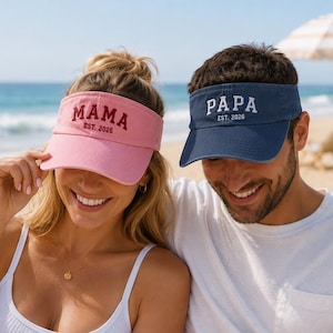 May include: A light pink visor with the embroidered text MAMA EST. 2026 in dark red and a navy blue visor with the embroidered text PAPA EST. 2026 in white. Both visors feature a curved brim and an adjustable strap design for headwear.