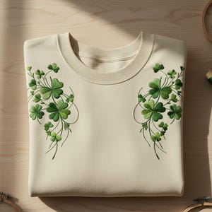 May include: Cream-colored sweatshirt with embroidered green shamrock designs on the front. The shamrocks are in varying shades of green, creating a detailed and textured appearance. The sweatshirt is folded neatly, showcasing the intricate embroidery.
