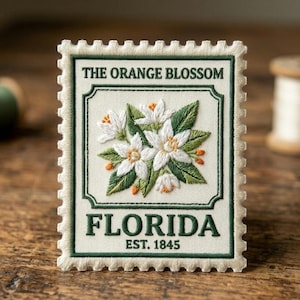 May include: Embroidered patch in the shape of a postage stamp. The design features white orange blossoms with green leaves and orange accents. The text "THE ORANGE BLOSSOM" and "FLORIDA EST. 1845" are embroidered in green.