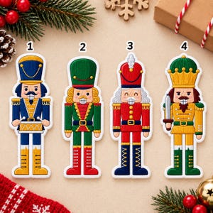 Könnte beinhalten: Vier gestickte Nussknacker-Ornamente, nummeriert von 1 bis 4. Nummer 1 ist blau und gold mit einer Trommel. Nummer 2 ist grün und rot mit Goldbesatz. Nummer 3 ist rot und blau mit einer weißen Feder. Nummer 4 ist gold und grün mit einem Schwert.