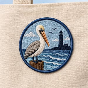 May include: Embroidered patch featuring a pelican perched on a wooden piling, with a lighthouse in the background. The patch has a blue border and depicts a coastal scene with waves, clouds, and birds. The patch is attached to a beige tote bag.